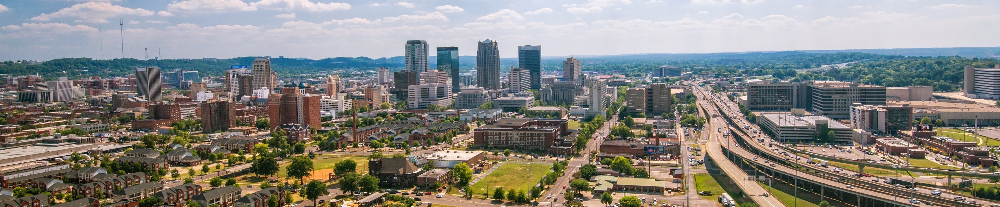 A wide view of the City of Birmingham, with smaller buildings in the foreground, roads and highways to the right, taller buildings in the left and background, and the blue sky above it all.