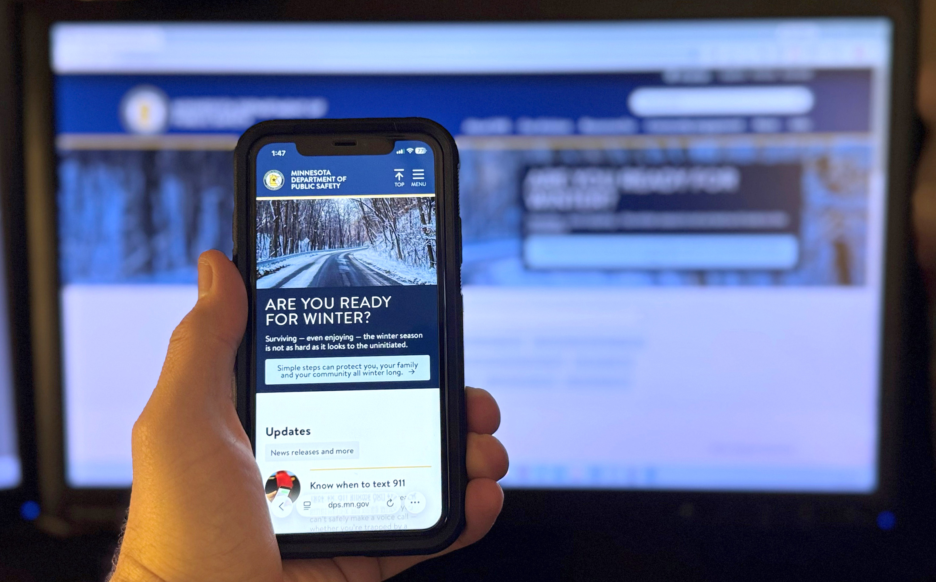 The DPS website homepage displayed on a smartphone being held up in front of a computer monitor that is also displaying the website homepage.