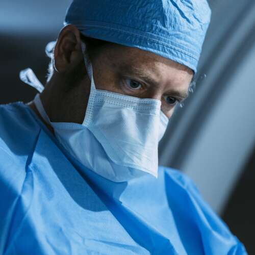 A doctor's face in blue scrubs, mask, and hat during a surgery