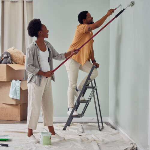 Couple paints a wall with painting tools and ladder.