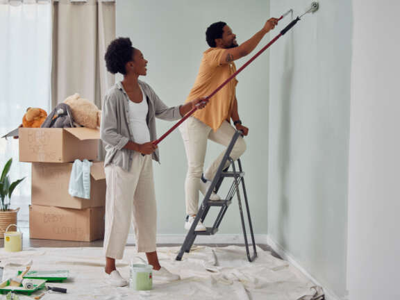 Couple paints a wall with painting tools and ladder.