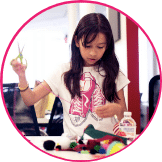 A young girl in a t-shirt with a sneakers print uses scissors to cut crafts at a table with various art supplies and a bottle of glue, sparking creativity that could be nurtured through entrepreneurship education.