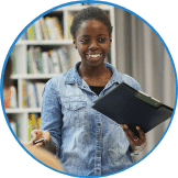 A person in a denim shirt is holding a clipboard and a pen, standing in front of bookshelves, illustrating the hands-on approach of youth entrepreneurship education.