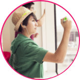 A young woman in a social entrepreneurship program, with a straw hat writing on a clear board, her reflection visible, in a brightly lit room.