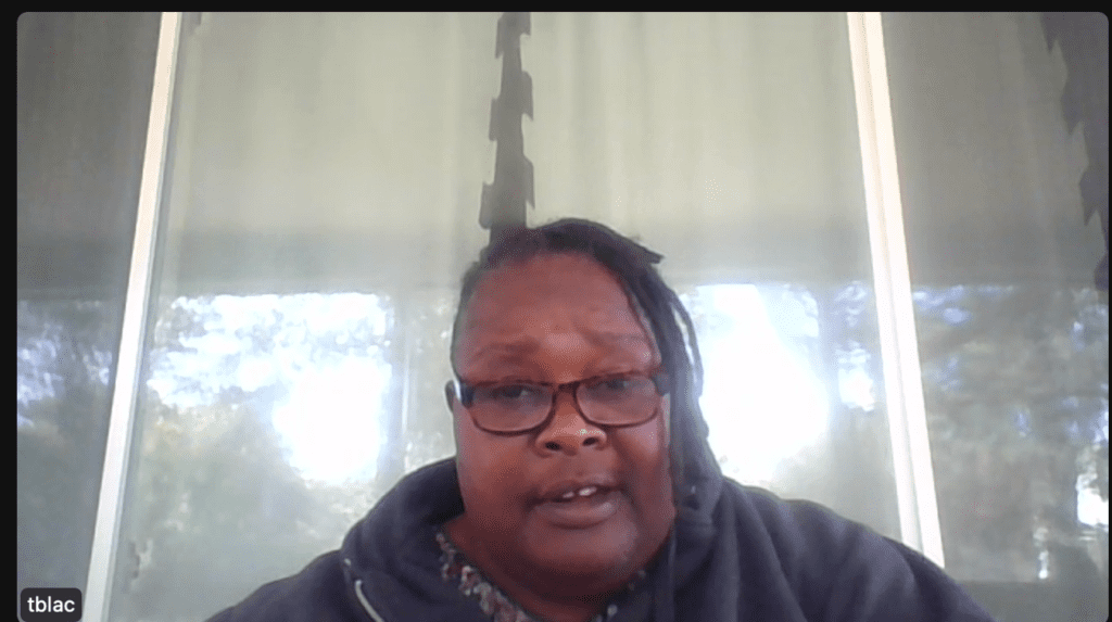 A person with glasses and dreadlocks is seated in front of a window with partially drawn blinds, wearing a dark hoodie, as they delve into youth entrepreneurship education.