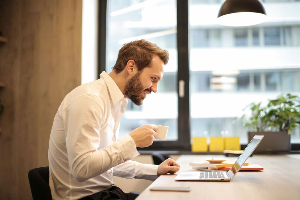 Man sitting at a modern office desk, smiling while reviewing his budget on a laptop using a calendar-based budgeting app, with a cup of coffee in hand.