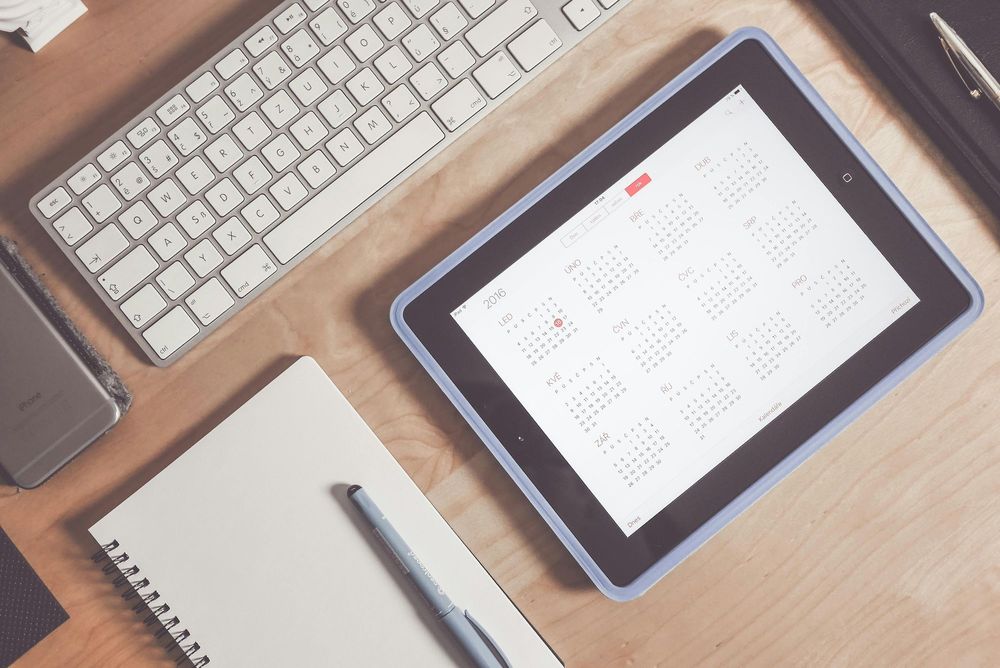 A tablet displaying a digital calendar on a wooden desk beside a keyboard, notebook, and pen — representing calendar-based budgeting and financial planning.
