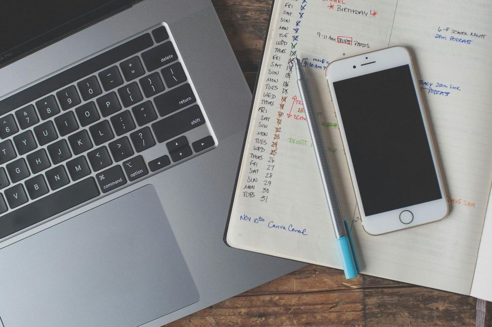 A laptop, smartphone, and handwritten calendar planner on a wooden desk — representing a calendar budgeting app setup for organizing expenses and tracking cash flow.