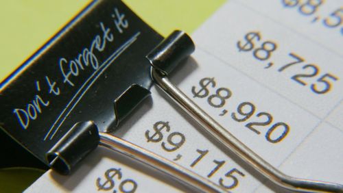 Close-up of a black binder clip labeled “Don’t forget it” holding a financial document showing dollar amounts such as $8,725 and $9,115, symbolizing budgeting or expense tracking.