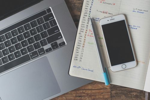 A laptop, smartphone, and handwritten calendar planner on a wooden desk — representing a calendar budgeting app setup for organizing expenses and tracking cash flow.