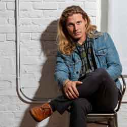 A casually dressed person with long blond hair sits on a chair against a white brick wall, wearing a denim jacket, dark pants, and brown shoes.