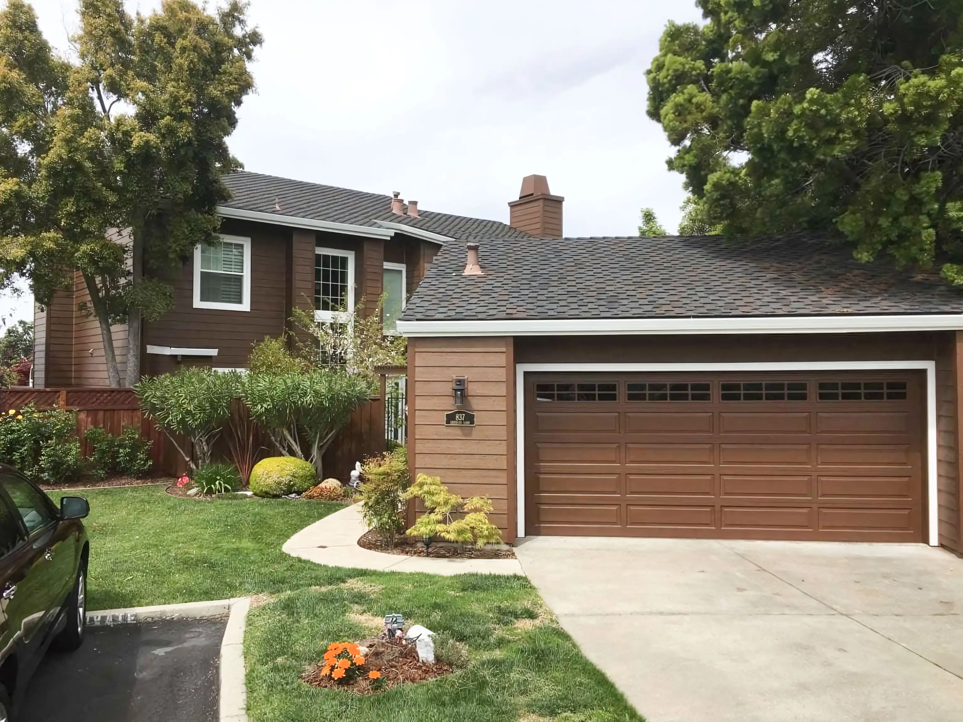  A two-story house with brown siding and white trim, a well-maintained lawn, and a brown garage door surrounded by trees and shrubs.