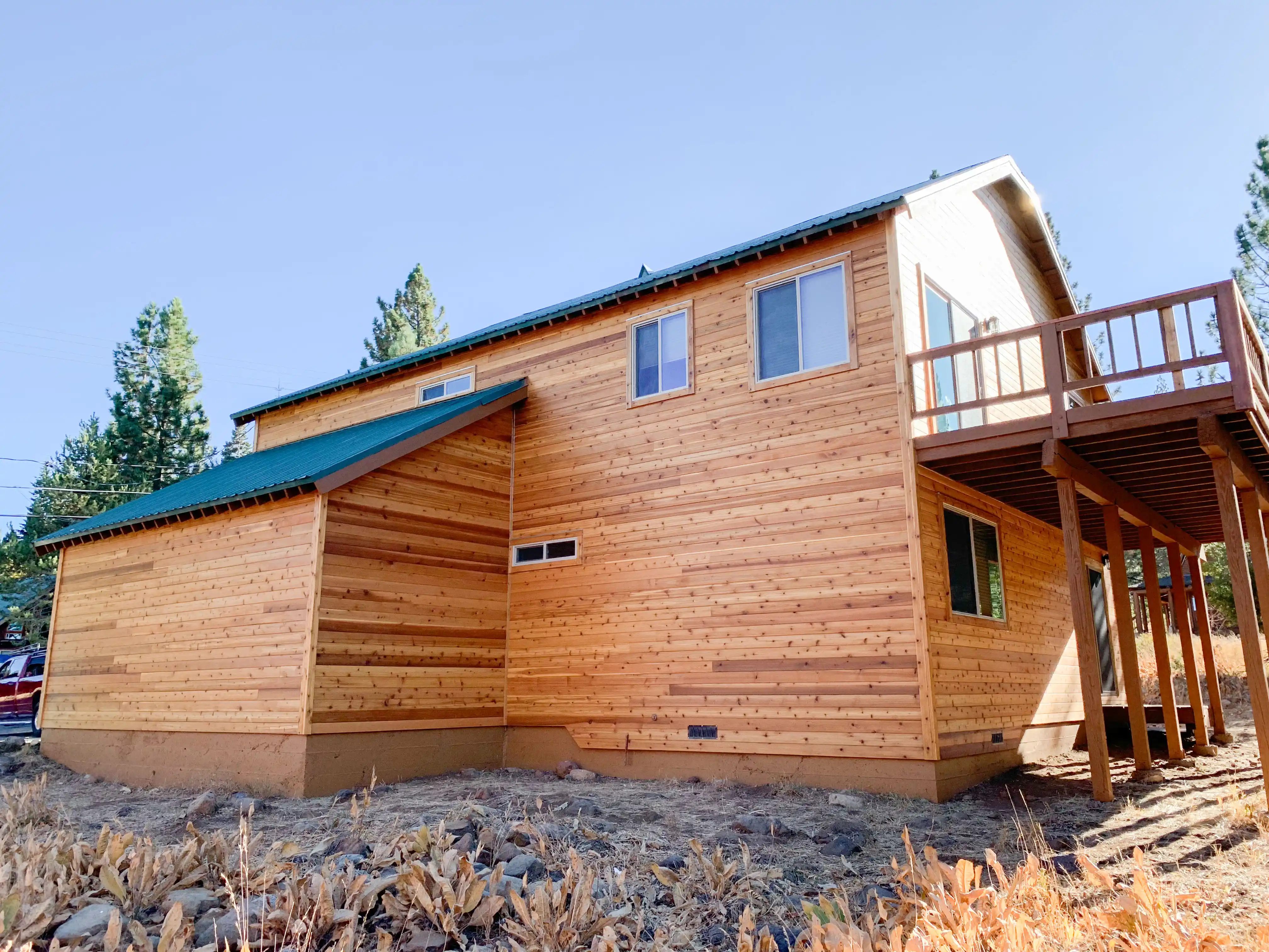 A two-story house with a newly constructed wooden exterior and a green roof. Elevated wooden deck on the right side, surrounded by dry grass and clear sky. A two-story house with a newly constructed wooden exterior and a green roof. Elevated wooden deck on the right side, surrounded by dry grass and clear sky.