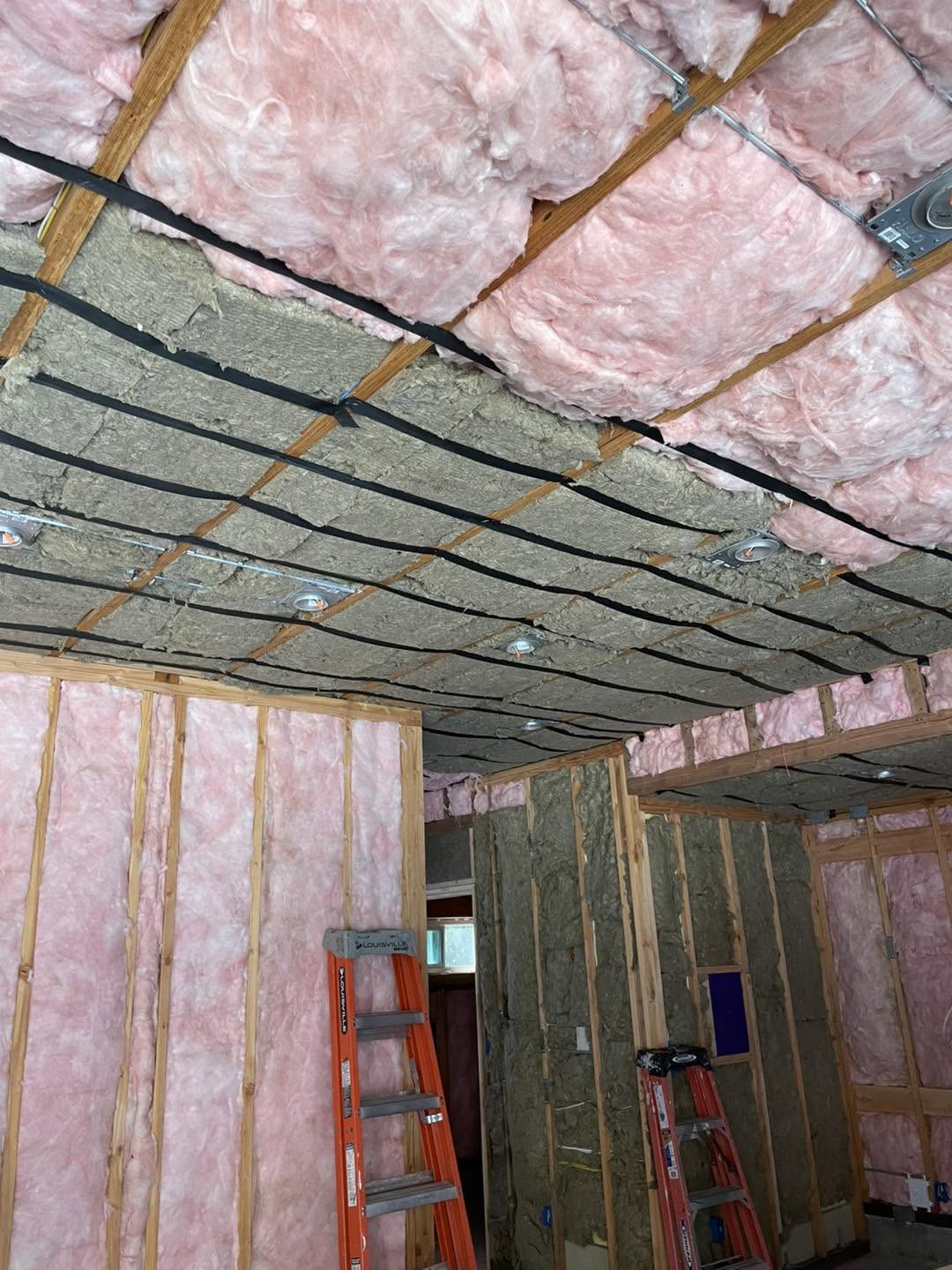 Exposed wood framing with pink fiberglass insulation in the walls and ceiling and gray mineral wool insulation installed between ceiling joists.