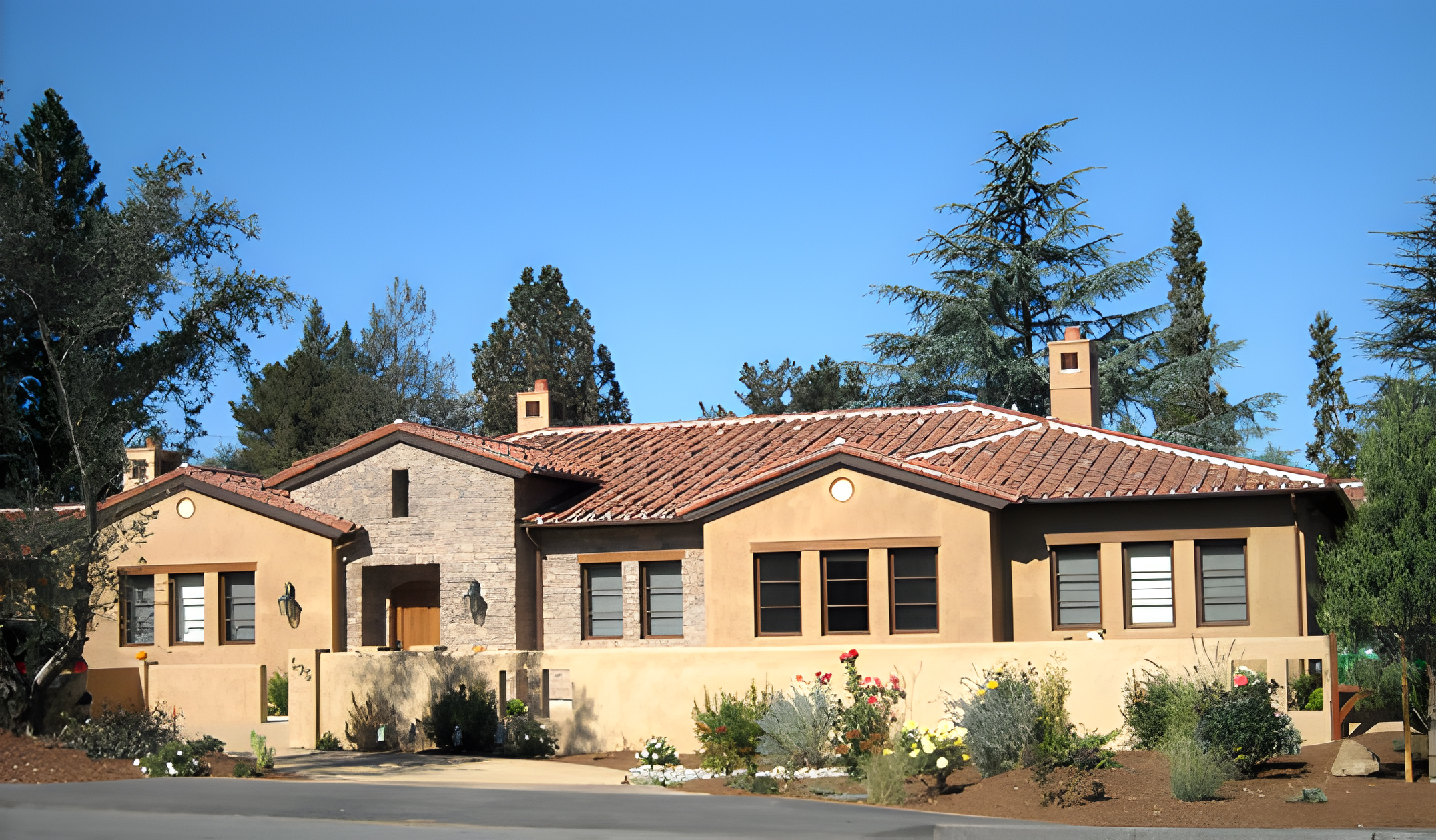  A tan stucco house with a terracotta roof, surrounded by trees and landscaped gardens, under a clear blue sky.