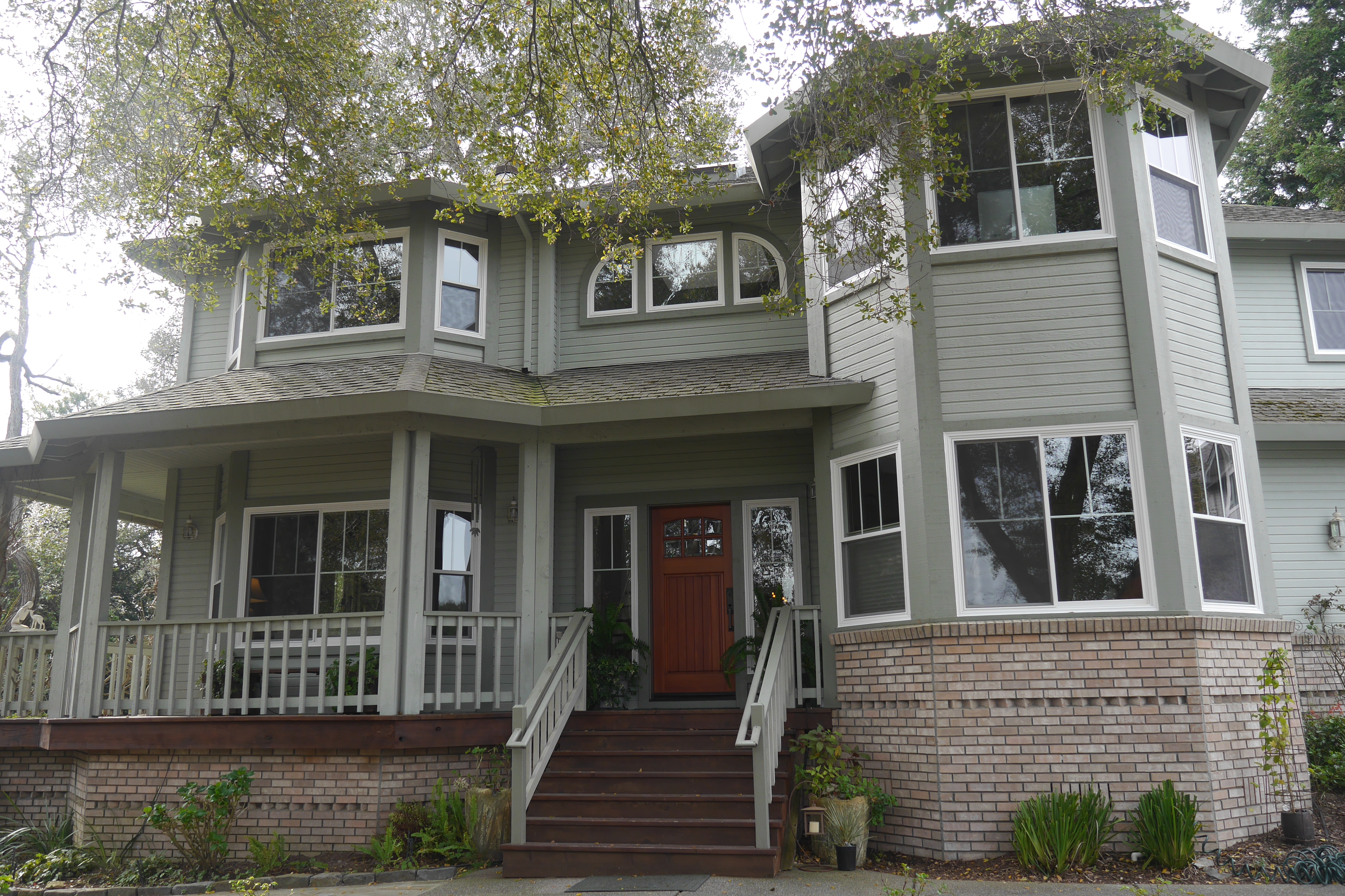  A two-story house with a gray exterior, large windows, and a plant-lined porch, partially shaded by trees.