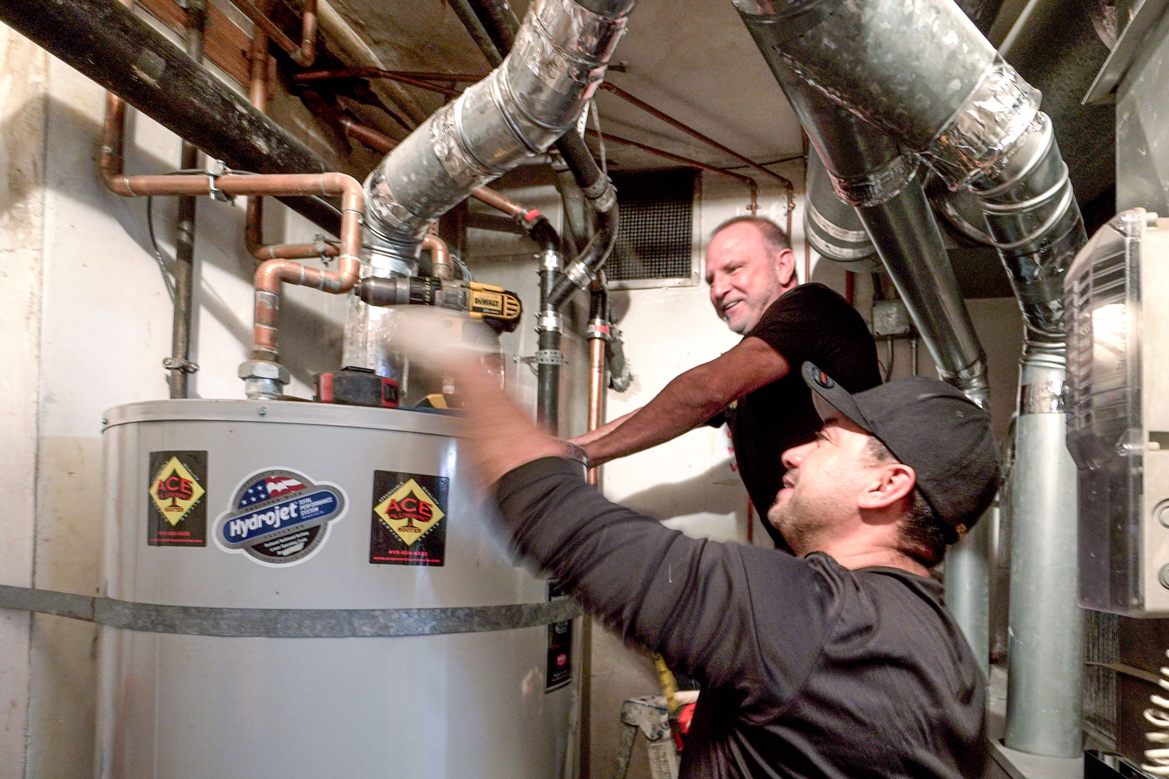 Two plumbers working on a water heater in a basement with pipes and ducts overhead. Two plumbers working on a water heater in a basement with pipes and ducts overhead.
