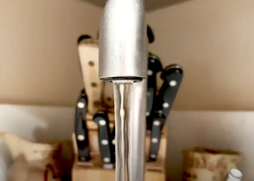 Close-up of water flowing from a kitchen faucet with a knife block in the background. Close-up of water flowing from a kitchen faucet with a knife block in the background.