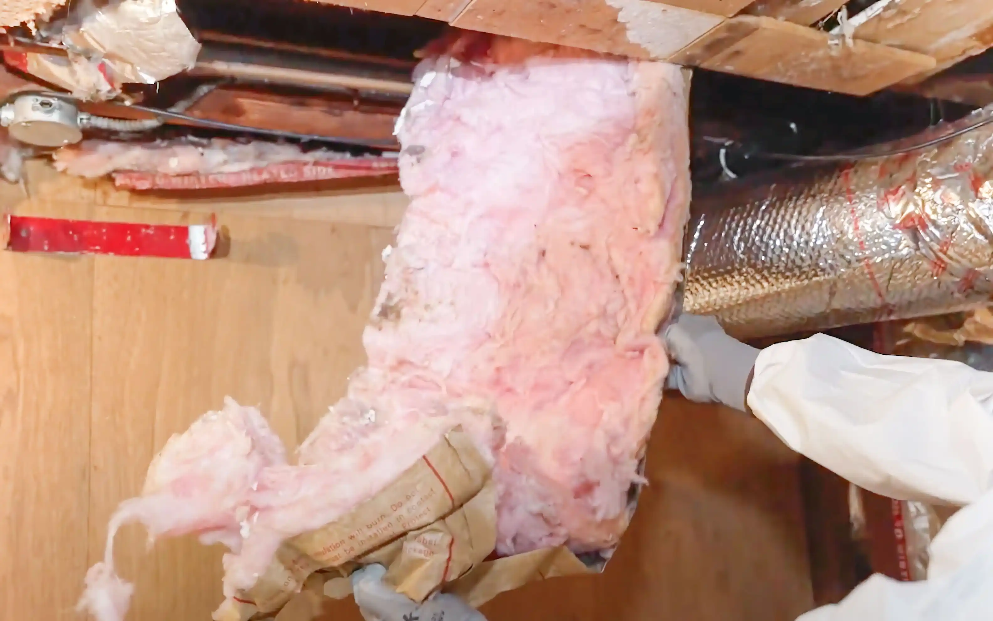 Worker in protective gear handling pink insulation material near ductwork in an unfinished space. Worker in protective gear handling pink insulation material near ductwork in an unfinished space.