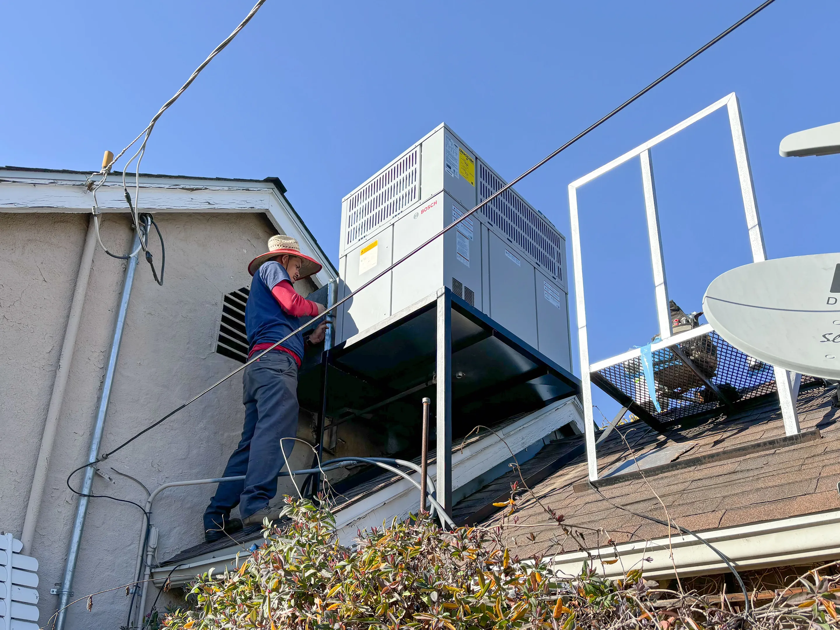 Technician on a roof examining an AC unit with a straw hat, next to a satellite dish, under a clear blue sky. Technician on a roof examining an AC unit with a straw hat, next to a satellite dish, under a clear blue sky.