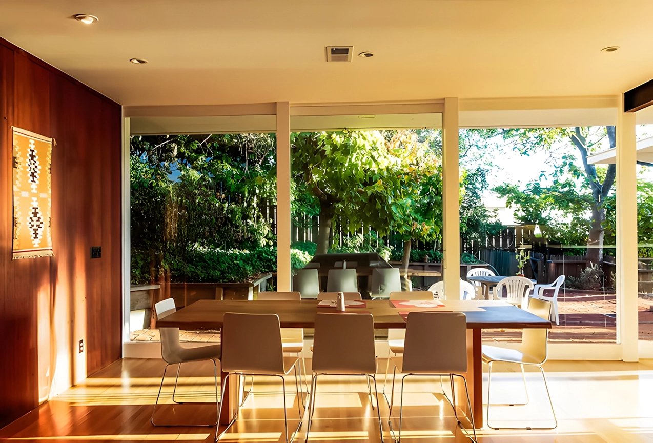 Dining room with large windows, wooden table, white chairs, and view of garden outside. Dining room with large windows, wooden table, white chairs, and view of garden outside.