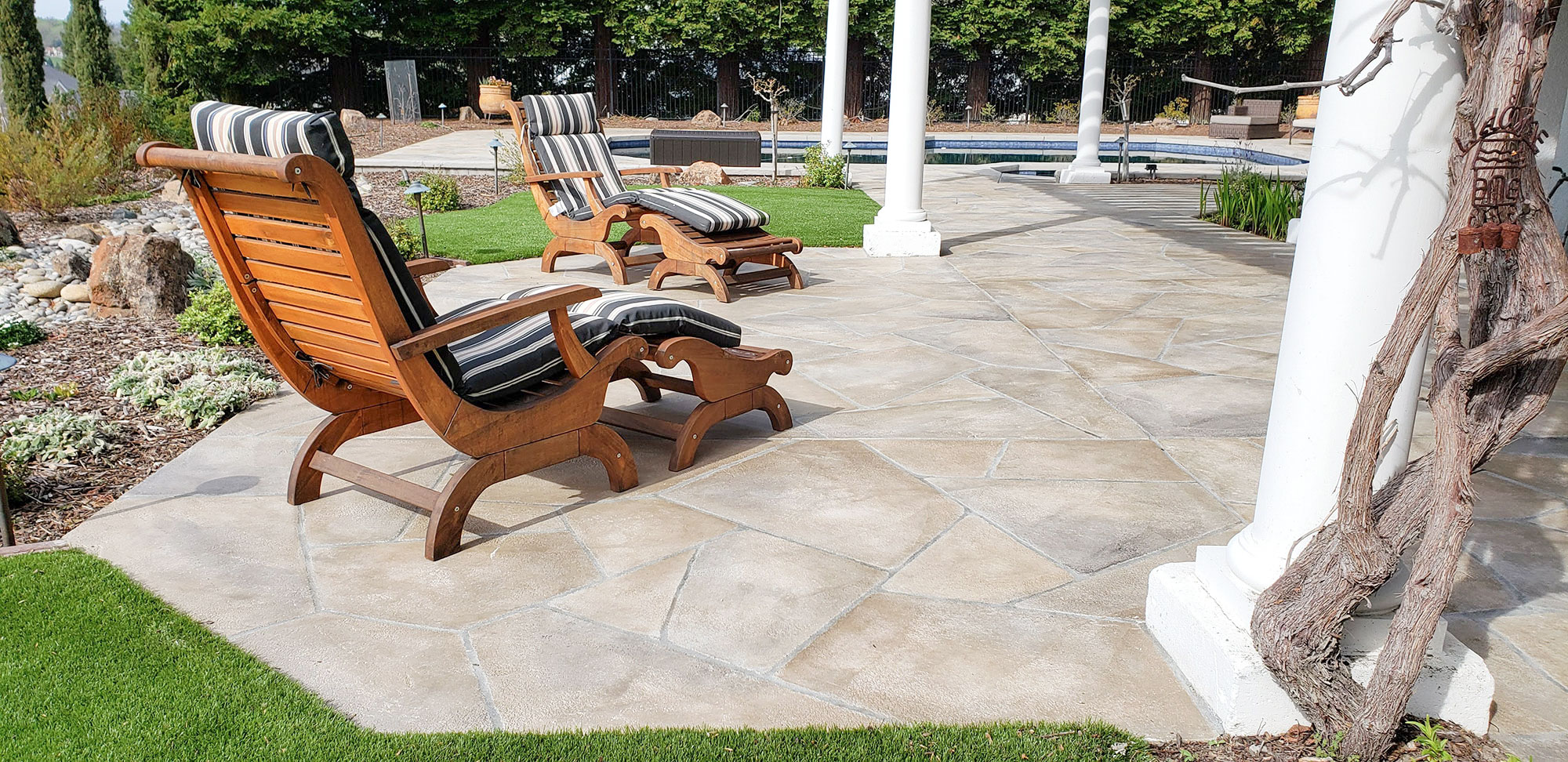  Outdoor wooden recliners with striped cushions on a stone patio, near columns and landscaped greenery.