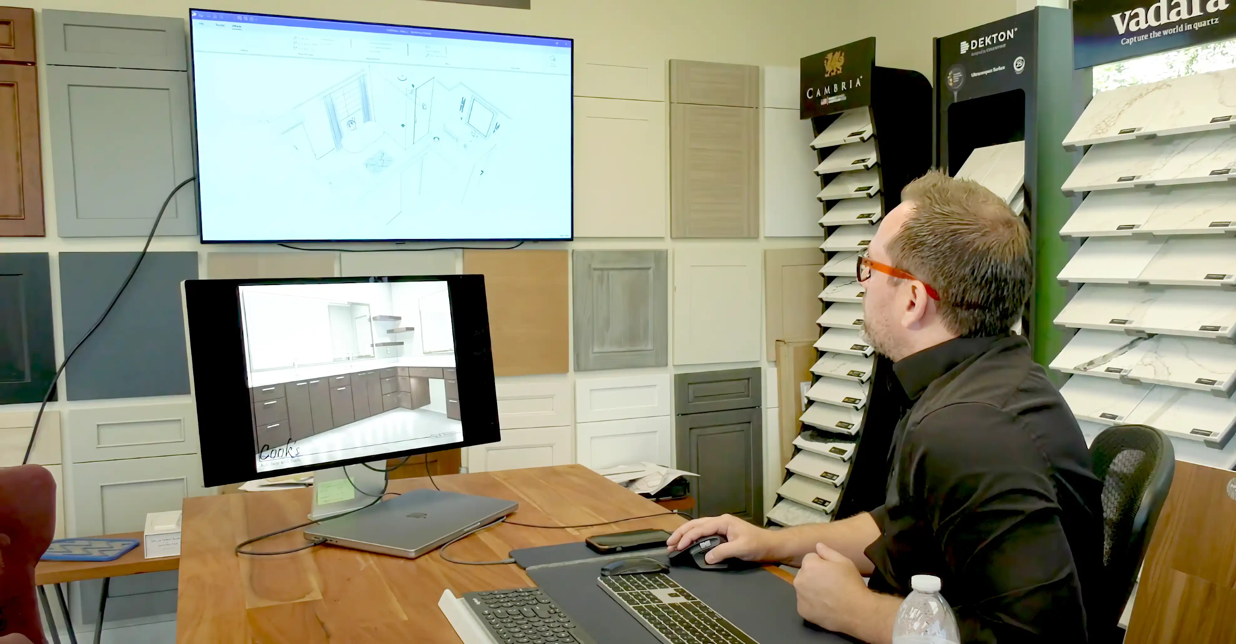 A person reviews kitchen design plans on two monitors, surrounded by cabinet samples and countertop displays in an office setting. A person reviews kitchen design plans on two monitors, surrounded by cabinet samples and countertop displays in an office setting.