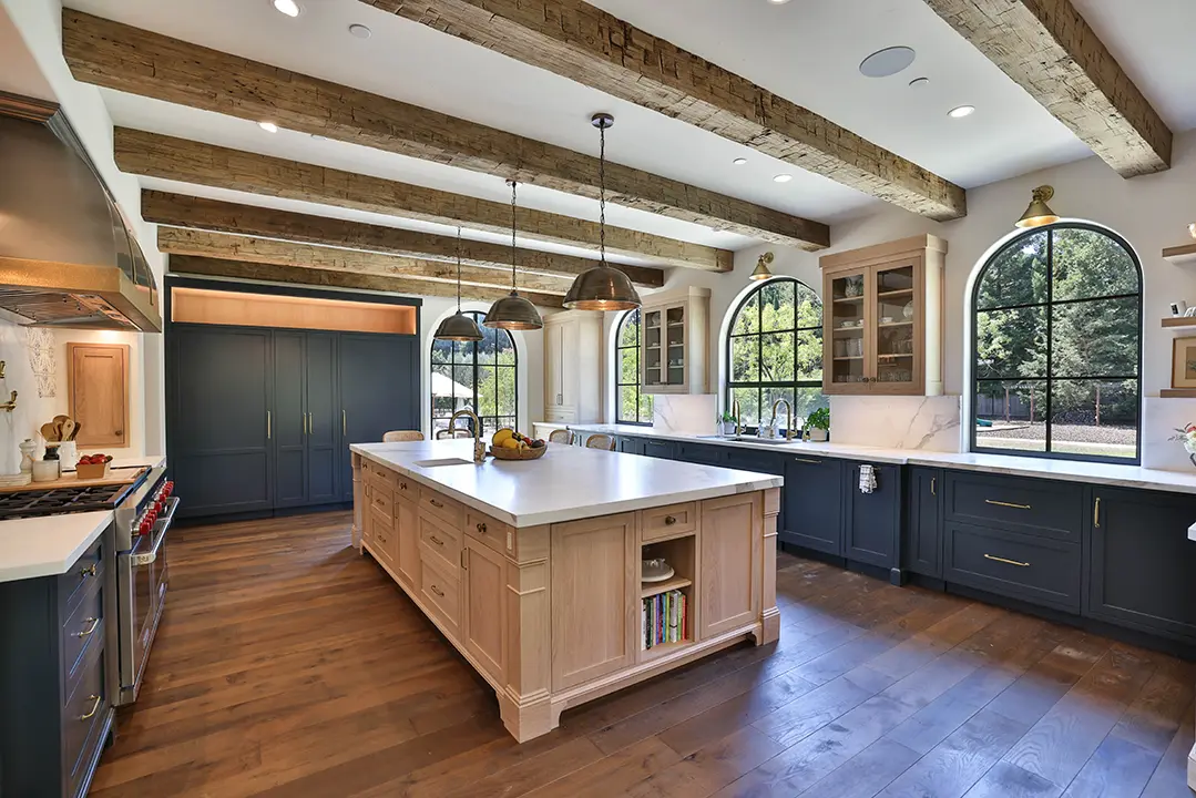  Spacious kitchen with wooden beams, large island, dark cabinetry, and arched windows overlooking greenery.