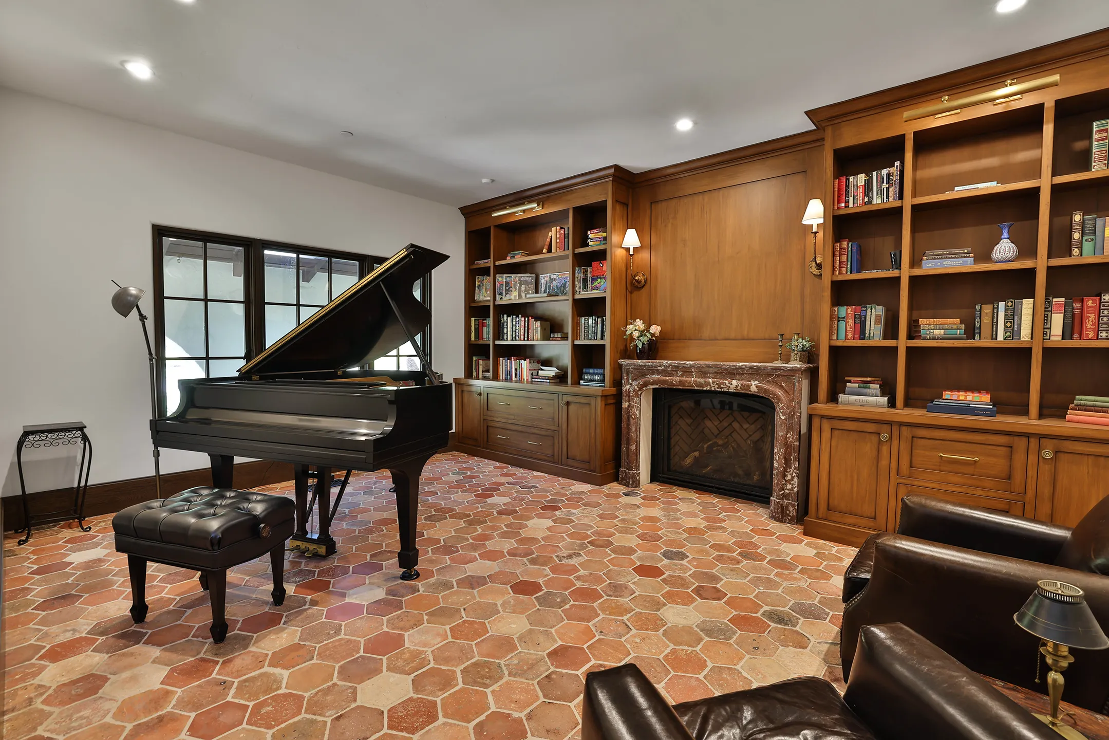 Room with a grand piano on a terracotta floor, wooden bookshelves, and a marble fireplace, illuminated by overhead lights. Room with a grand piano on a terracotta floor, wooden bookshelves, and a marble fireplace, illuminated by overhead lights.