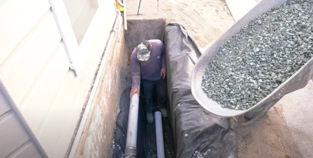 Worker in trench adjusting drainage pipe next to a pile of gravel in a wheelbarrow, beside a building. Worker in trench adjusting drainage pipe next to a pile of gravel in a wheelbarrow, beside a building.