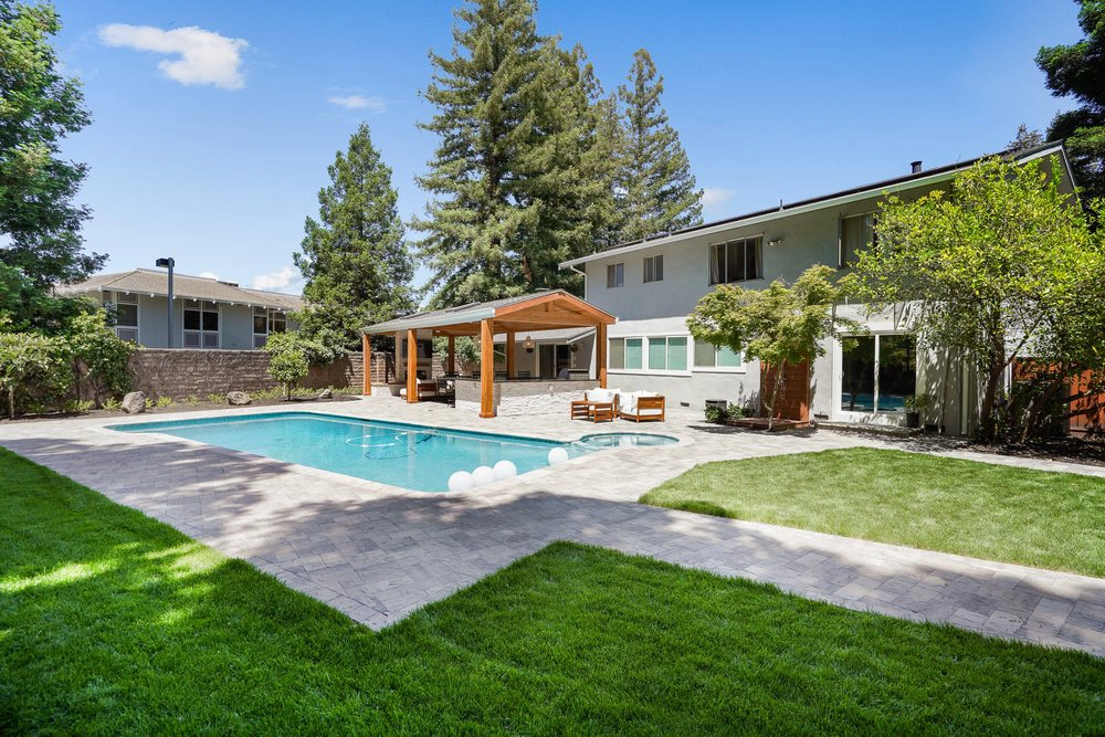 Spacious backyard with a swimming pool, wooden pergola, and outdoor seating. Surrounded by lush grass and mature trees, adjacent to a two-story house. Spacious backyard with a swimming pool, wooden pergola, and outdoor seating. Surrounded by lush grass and mature trees, adjacent to a two-story house.