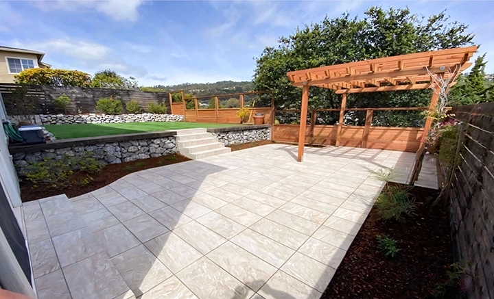Spacious patio with polished tiles, a wooden pergola, steps leading to a grassy area, and surrounding greenery. Spacious patio with polished tiles, a wooden pergola, steps leading to a grassy area, and surrounding greenery.