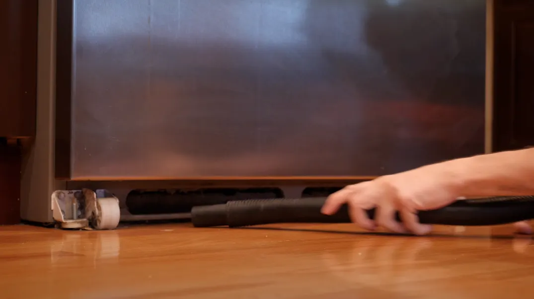 Close-up of a hand using a vacuum hose to clean under the bottom edge of a refrigerator on a wooden floor.