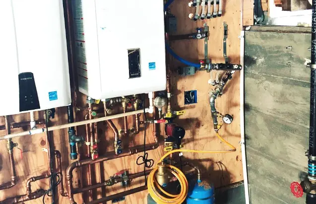 A detailed view of a plumbing installation with pipes, valves, gauges, and two large white units mounted on a wooden wall panel. A detailed view of a plumbing installation with pipes, valves, gauges, and two large white units mounted on a wooden wall panel.
