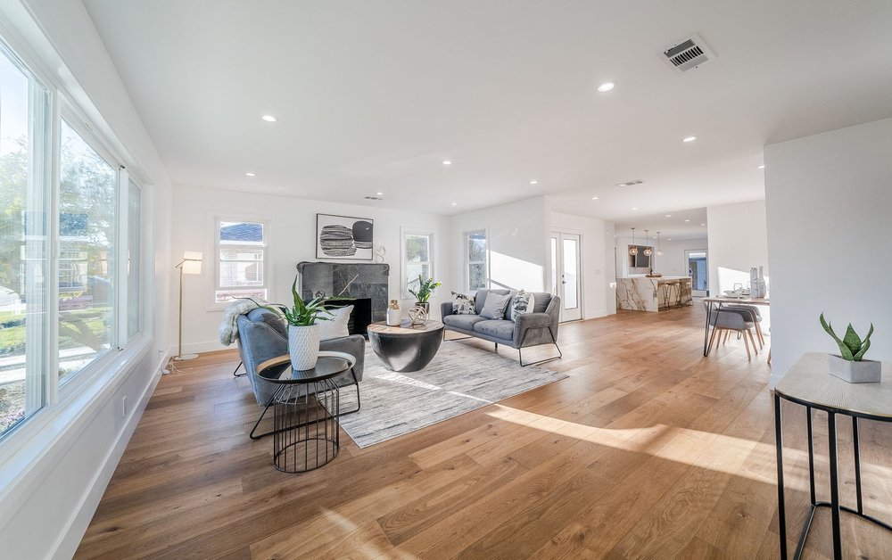 Open living room with large windows, wooden floor, gray furniture, modern art, and a view into the connected kitchen area. Open living room with large windows, wooden floor, gray furniture, modern art, and a view into the connected kitchen area.