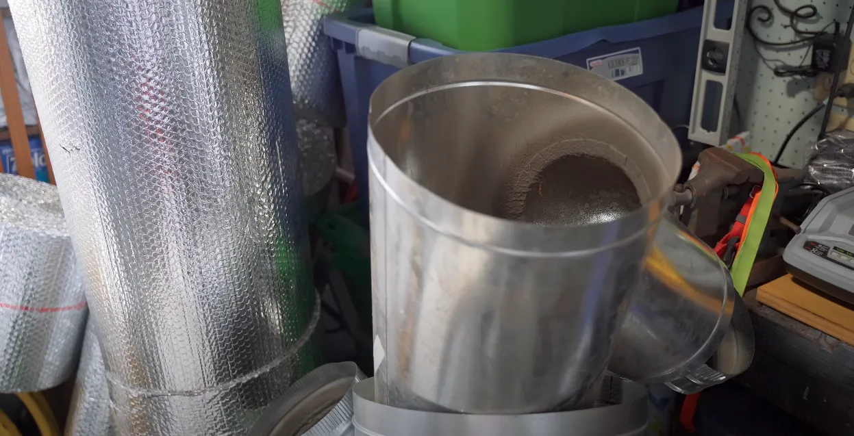A shiny steel duct section with insulating material nearby, placed in a cluttered workshop setting. A shiny steel duct section with insulating material nearby, placed in a cluttered workshop setting.