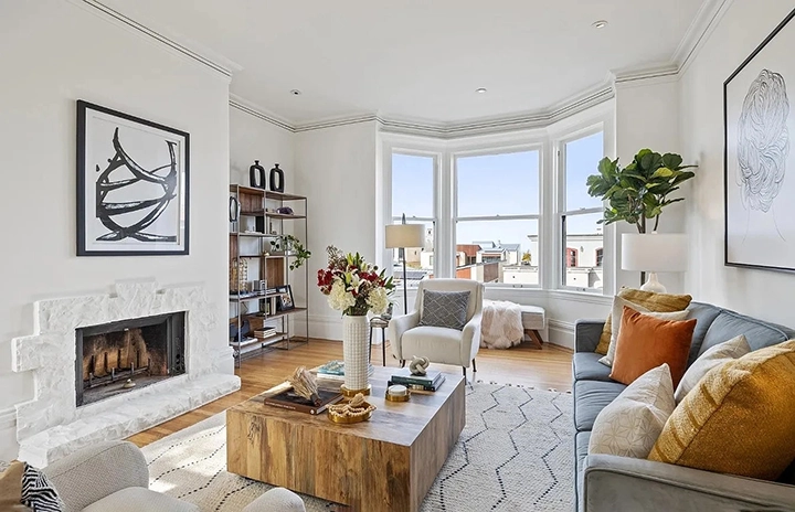A bright living room with a fireplace, large windows, a wooden coffee table, and cushioned seating with pillows. A bookshelf and plants add decor. A bright living room with a fireplace, large windows, a wooden coffee table, and cushioned seating with pillows. A bookshelf and plants add decor.