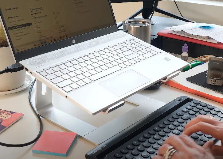 A person types on a keyboard at a desk with a laptop on a stand, surrounded by notes, a coffee cup, and office supplies. A person types on a keyboard at a desk with a laptop on a stand, surrounded by notes, a coffee cup, and office supplies.