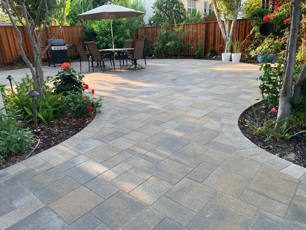 A well-maintained patio with interlocking pavers, surrounded by a garden with trees and flowers, and featuring a table with chairs and an umbrella. A well-maintained patio with interlocking pavers, surrounded by a garden with trees and flowers, and featuring a table with chairs and an umbrella.