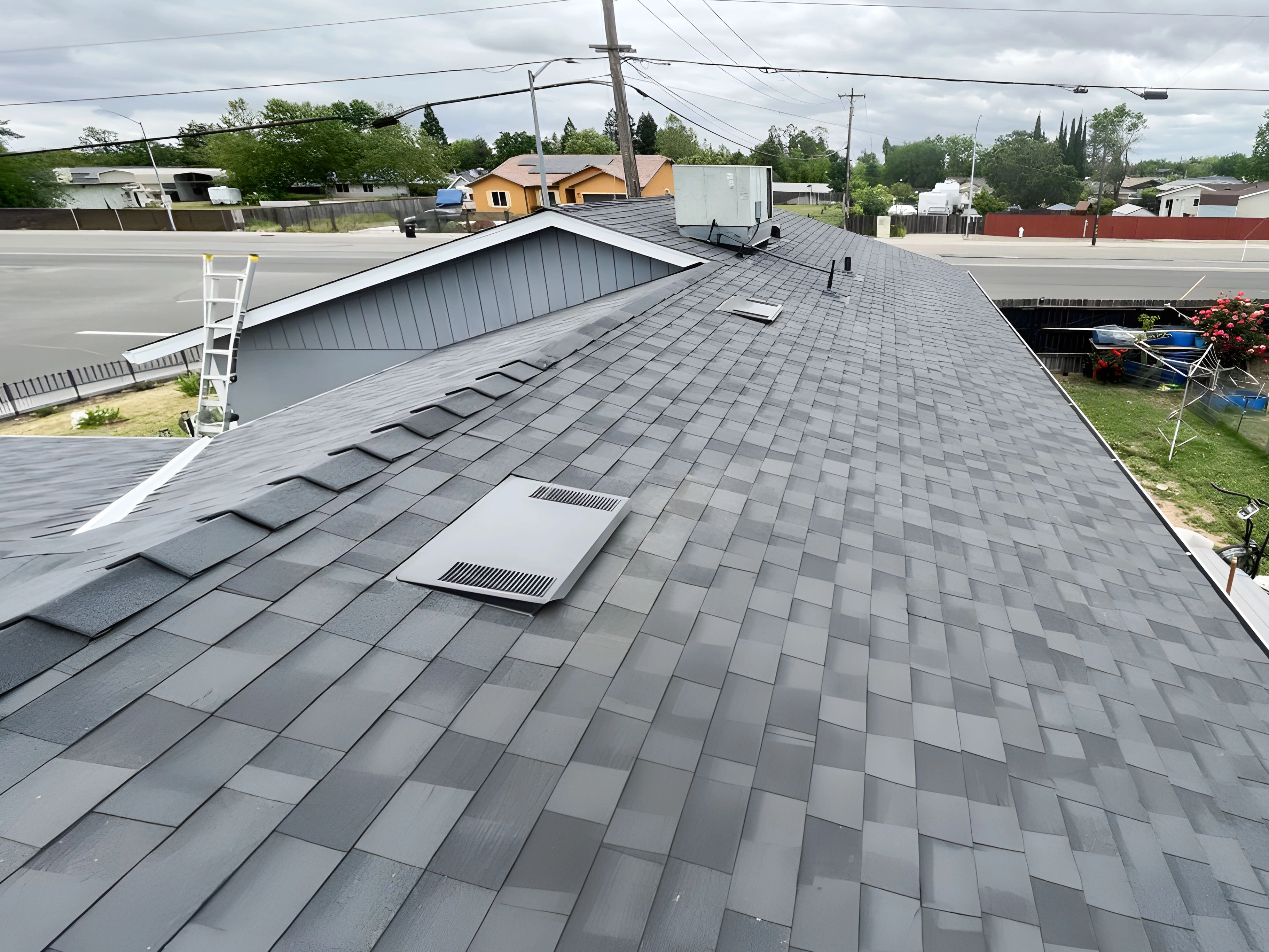 A rooftop with gray shingles, vents, and a ladder leading up to it. The surrounding area includes streets and residential homes. A rooftop with gray shingles, vents, and a ladder leading up to it. The surrounding area includes streets and residential homes.
