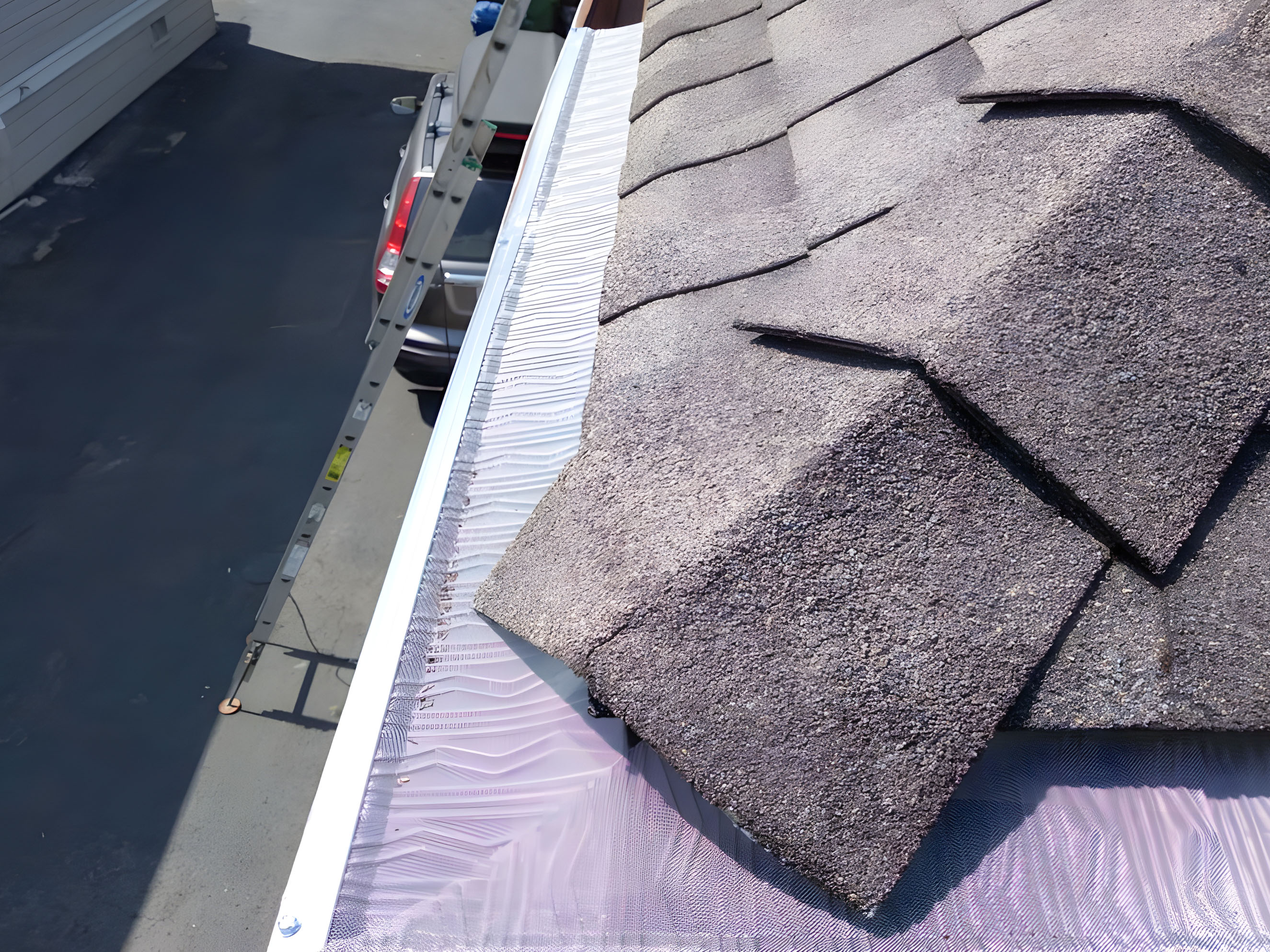 Close-up of shingles and gutter guard on a roof. A ladder leans against the building, and a vehicle is parked below on the driveway. Close-up of shingles and gutter guard on a roof. A ladder leans against the building, and a vehicle is parked below on the driveway.