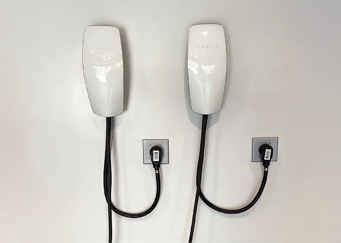 Two white electric vehicle chargers mounted on a wall, each with a cable plugged into a socket below. Two white electric vehicle chargers mounted on a wall, each with a cable plugged into a socket below.