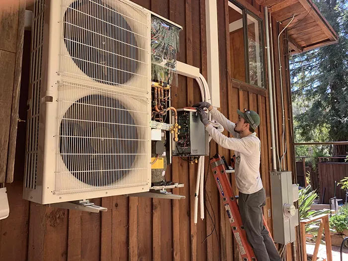  Technician on ladder working on a large air conditioning unit mounted on a wooden exterior wall, with tools and wires visible.