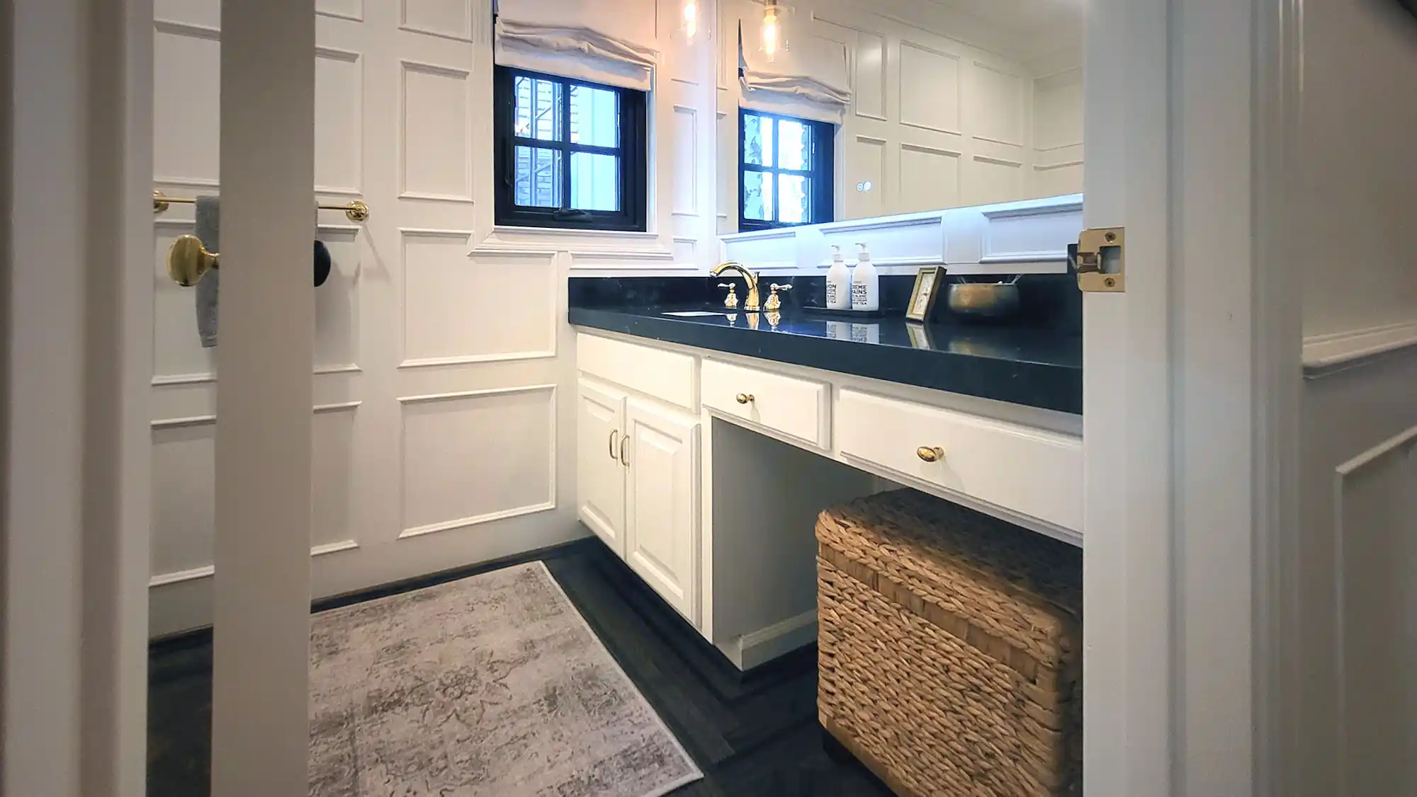 Bathroom with white paneled walls, black countertop, gold fixtures, a rectangular mirror, and a woven basket under the sink. Bathroom with white paneled walls, black countertop, gold fixtures, a rectangular mirror, and a woven basket under the sink.