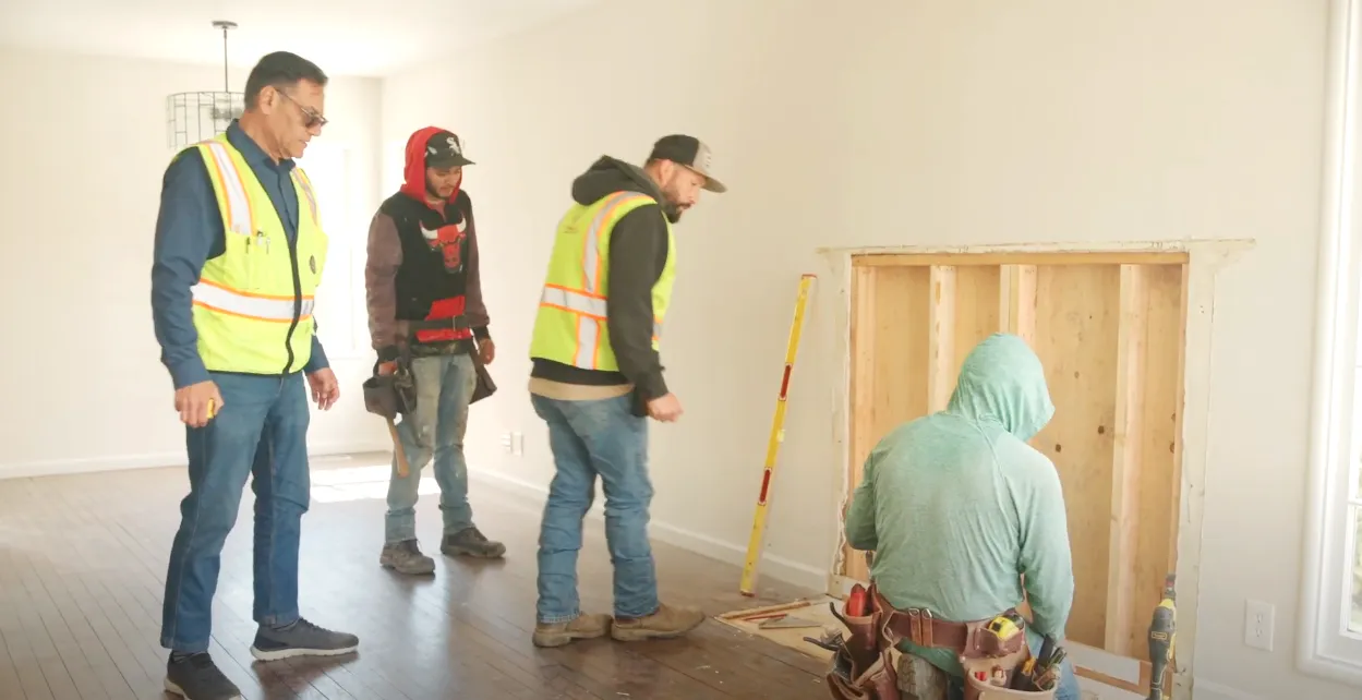 Four construction workers in safety gear examine wall framing in a bright room. A person kneels near the wall with tools close by. Four construction workers in safety gear examine wall framing in a bright room. A person kneels near the wall with tools close by.