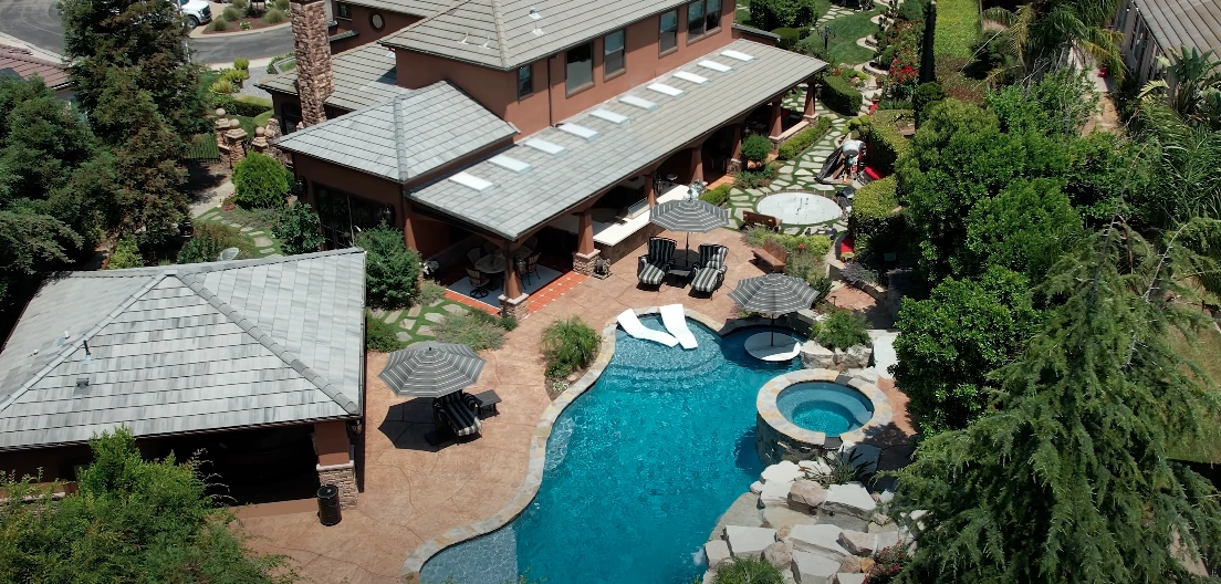 Aerial view of a landscaped backyard featuring a pool with surrounding patio furniture, umbrellas, and lush greenery.