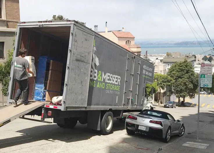 This image shows two professional movers from Robb and Messer Moving and Storage working together to transport a large wooden dresser. They are wearing company uniforms and using proper lifting techniques to ensure the safe handling of the furniture. The scene is set in a residential hallway, highlighting the movers' commitment to careful and efficient service during home relocations. Boxes and other packed items are visible in the background, emphasizing the comprehensive moving support provided by the team.