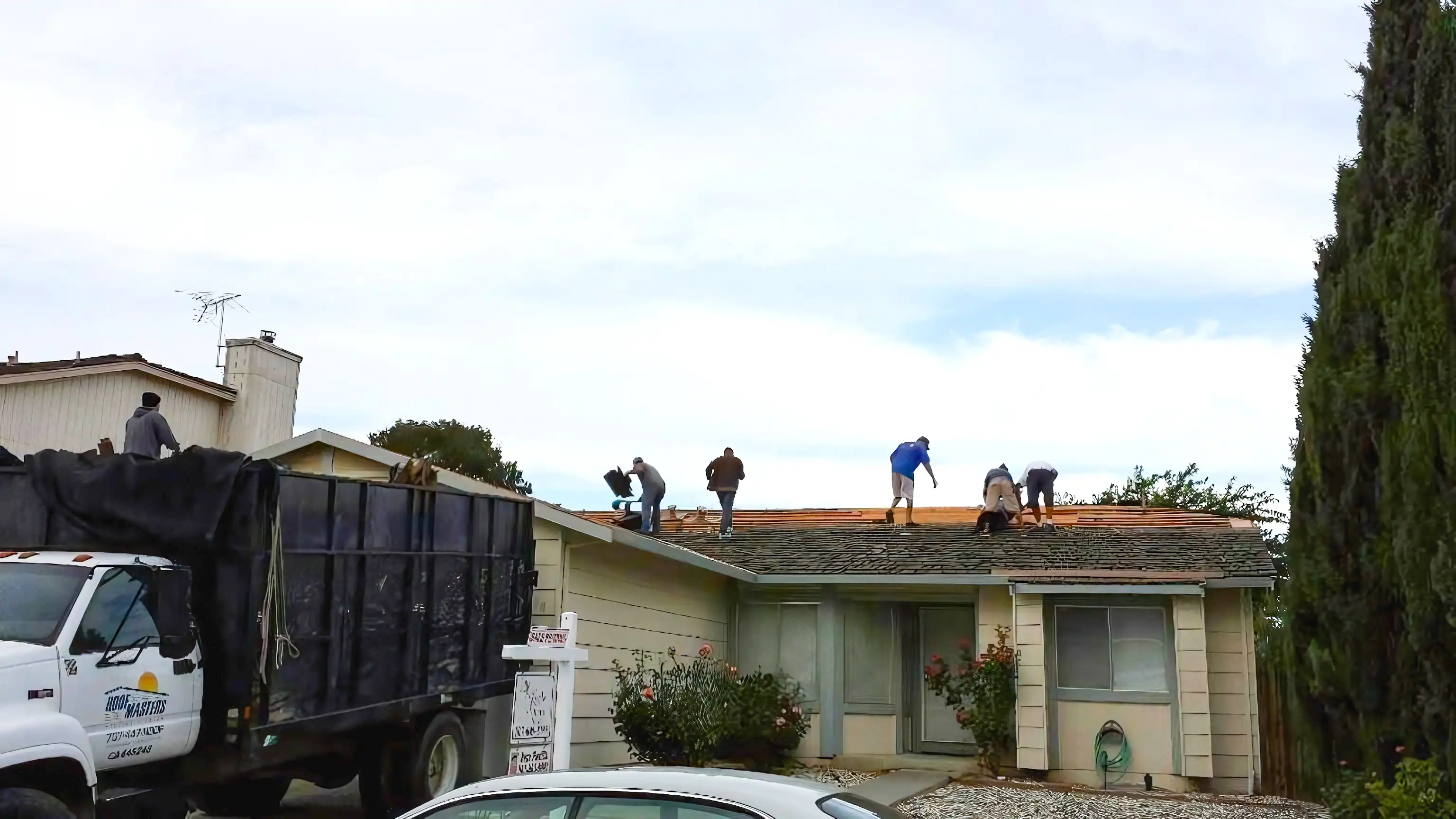  Workers on a roof replacing shingles; a ROOFMASTERS truck is parked nearby.