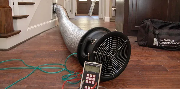 Air duct blower test setup on a wooden floor with a pressure gauge and hose extending into a hallway.