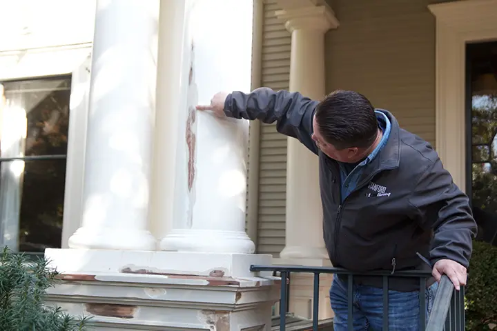 Person examining a weathered, peeling column on a porch, wearing a Stanford Painting, Inc. jacket. Person examining a weathered, peeling column on a porch, wearing a Stanford Painting, Inc. jacket.